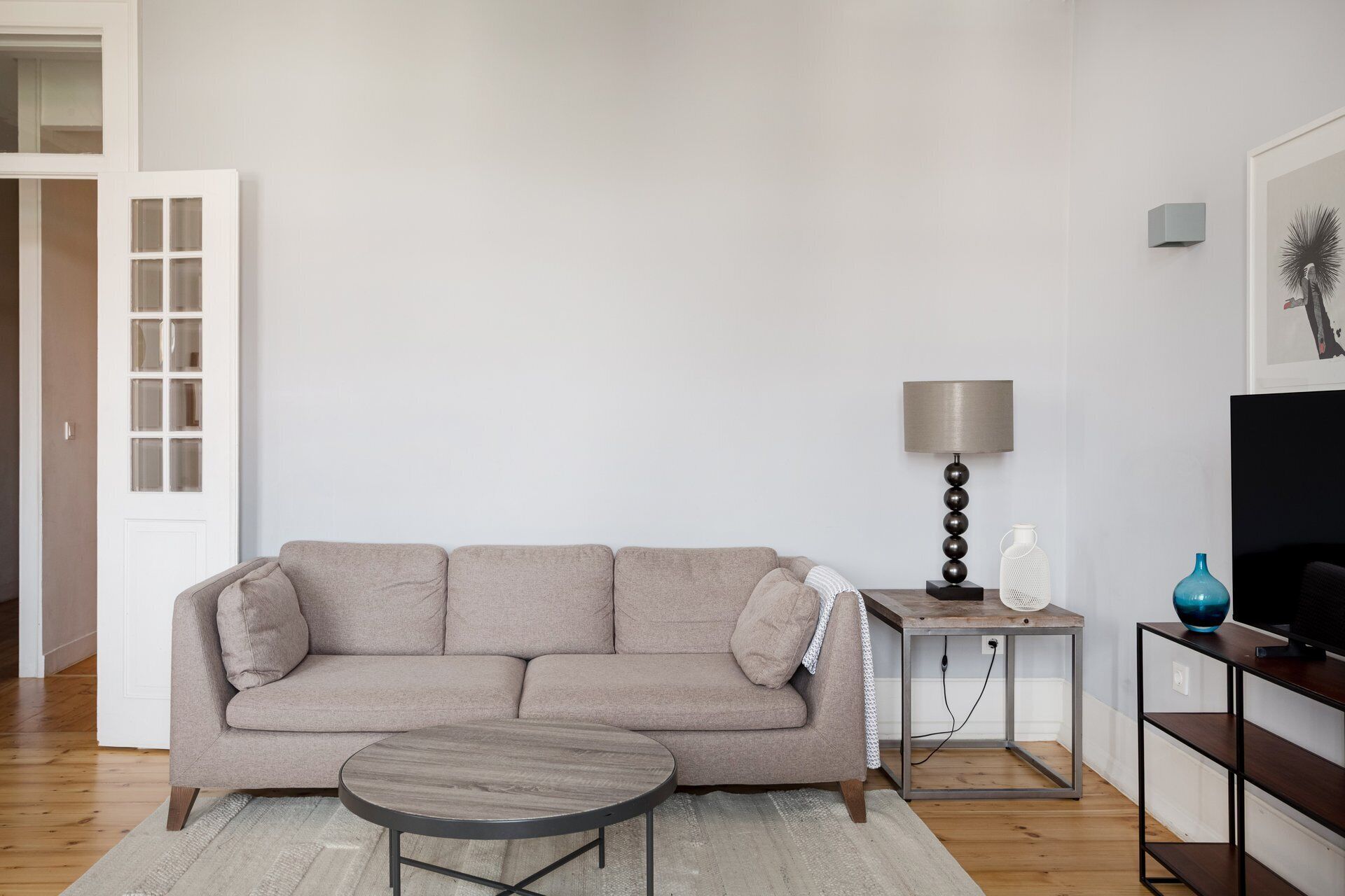 A living room with a sofa, a round coffee table, a lamp, a TV stand, and a door.