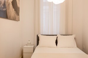 Un dormitorio con cama, mesita de noche, lámpara y ventana con cortinas translúcidas.