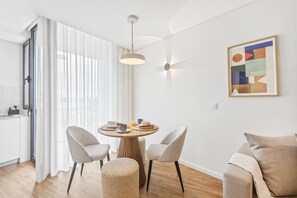 Ein modernes Wohnzimmer mit einem runden Esstisch, zwei Stühlen, einem Sofa, einer Küchenzeile und einem geometrischen Kunstwerk an der Wand.