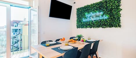 A dining area with a table set for four, a flat-screen TV mounted on the wall, and a neon sign.