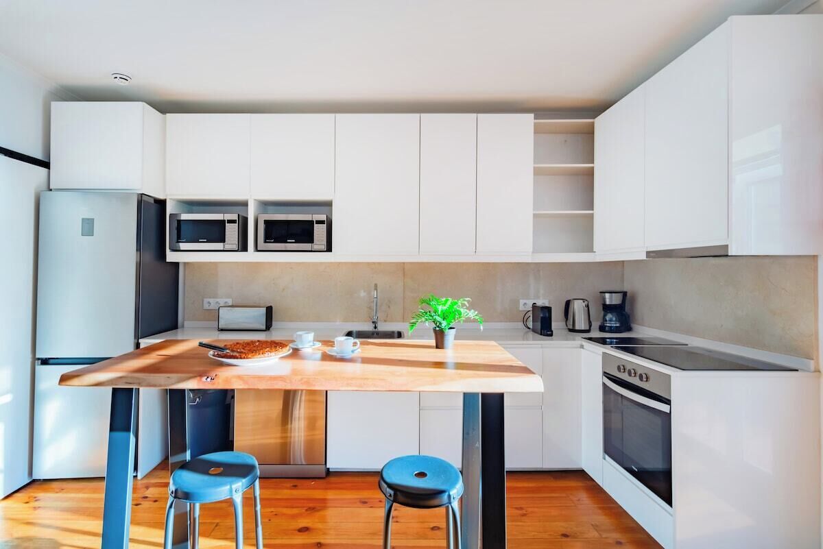Une cuisine moderne avec des meubles blancs, un îlot central en bois, deux tabourets, une machine à café et un four à micro-ondes.