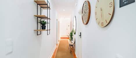 A hallway with a carpeted floor, a wooden bench, a plant, a wall clock, and a sign indicating room numbers.