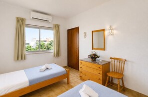 Ein Schlafzimmer mit Bett, Kommode, Stuhl, Klimaanlage, einem Fenster mit Vorhängen und einem Spiegel.
