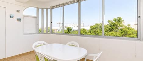 A room with a white table and chairs, large windows with blinds, and a view of greenery and buildings outside.