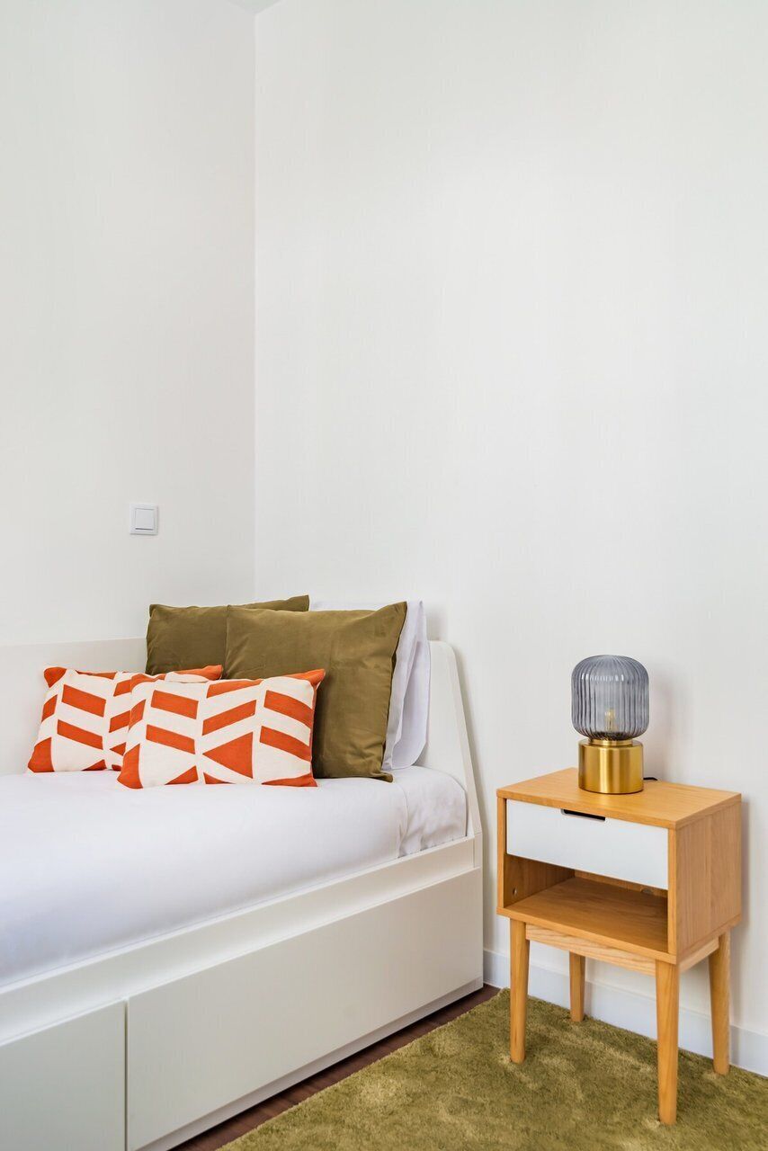 Un lit avec une literie blanche et des oreillers orange et marron, une table de chevet avec une lampe, et un tapis vert.