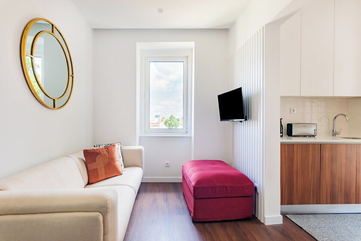 Un appartement moderne comprenant un canapé, un pouf rouge, un téléviseur et un coin cuisine.