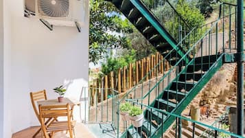 Eine Treppe mit grünem Metallgeländer führt zu einer erhöhten Ebene, Holzstühle und ein Tisch mit Topfpflanzen befinden sich auf einer sonnendurchfluteten Terrasse.