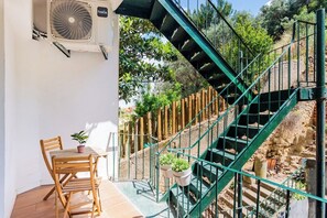 En trappe med grønt metalgelænder, der fører op til en højere etage, med træstole og et bord med plantede planter på en solbeskinnet terrasse.
