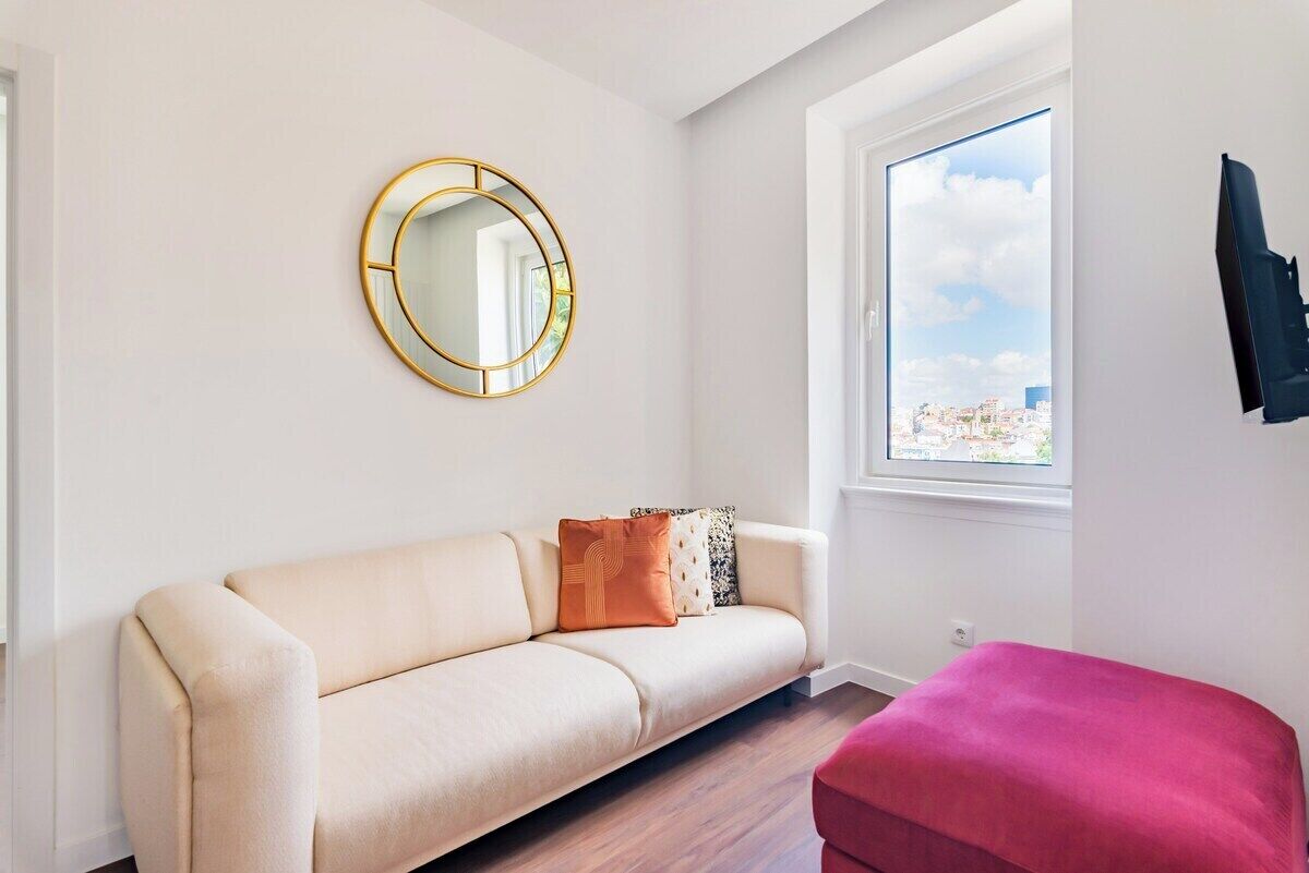 A room with a sofa, a round mirror, a window with a city view, and a pink ottoman.