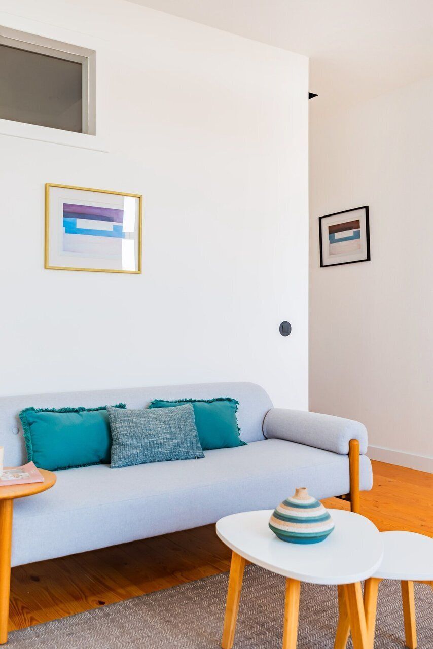 A modern living room with a light grey sofa, teal cushions, a small white table with a striped vase, and two white chairs.
