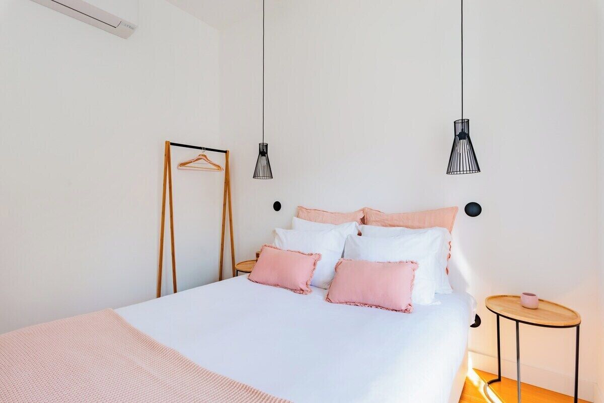 Uma cama bem arrumada em tons de branco e rosa, um porta-roupas de madeira e duas luzes pendentes.