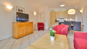Een moderne woonkamer met een televisie, een houten kast, een rode fauteuil, een eettafel en een keuken op de achtergrond.