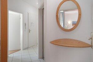 Un pasillo con una puerta de madera, una pared blanca con un espejo redondo y una estantería de madera.