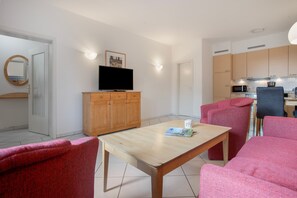 Una sala moderna con una mesa de madera, dos sillones rosados, un televisor sobre un mueble de madera y una zona de cocina con armarios y electrodomésticos.