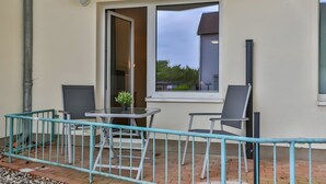 Een balkon met een tafel, twee stoelen, een plant in pot en uitzicht op een gebouw door een raam.