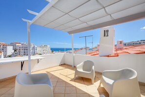 Eine Dachterrasse mit weißen Stühlen, einer weißen Decke und Blick auf Meer und Gebäude.