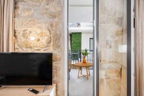 Quarto com parede de pedra, televisão sobre suporte de madeira, controle remoto e varanda com cadeira de vime e um vaso.