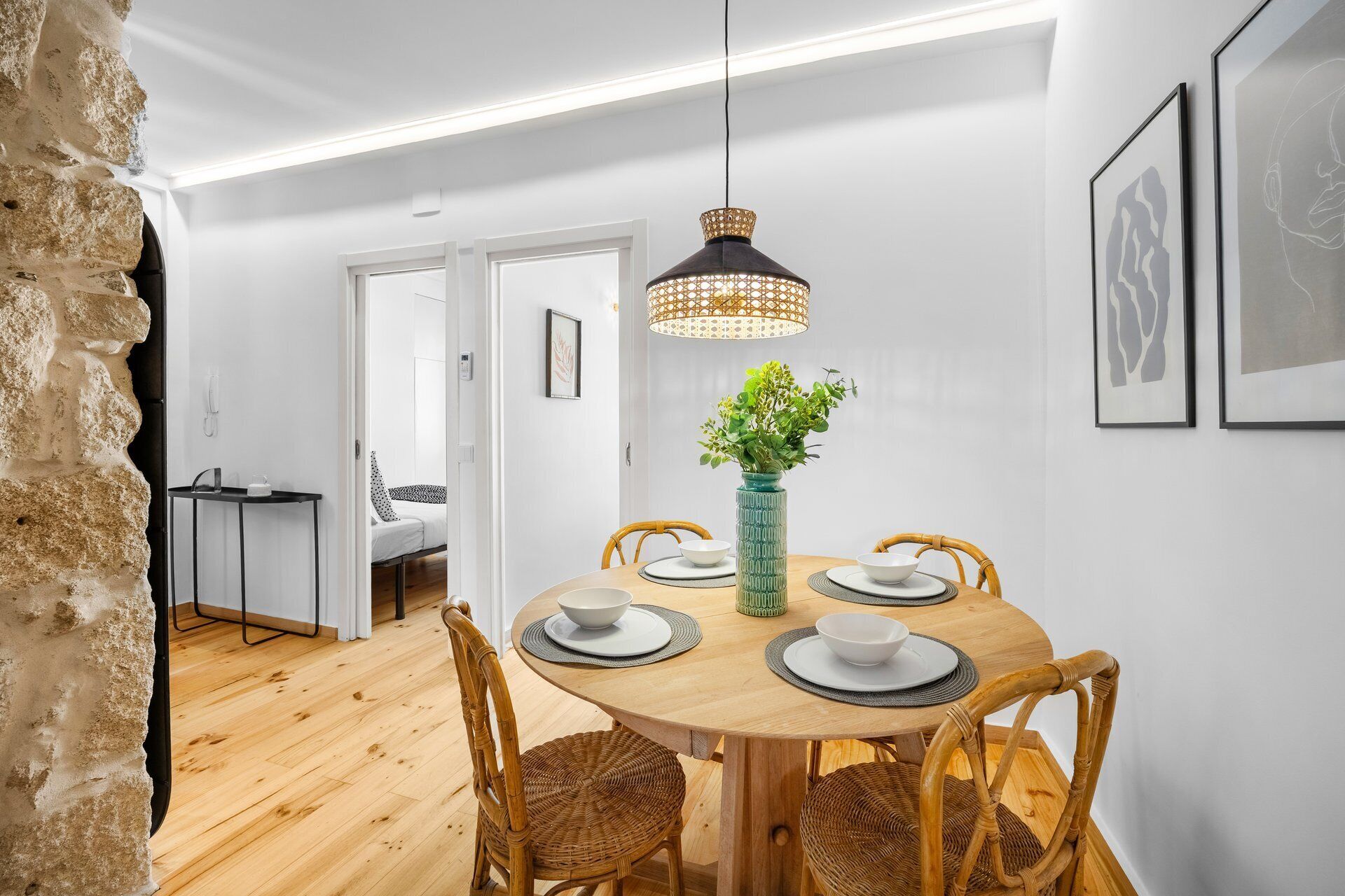 A dining area with a round wooden table, four wicker chairs, a green vase with flowers, and a hanging pendant light.