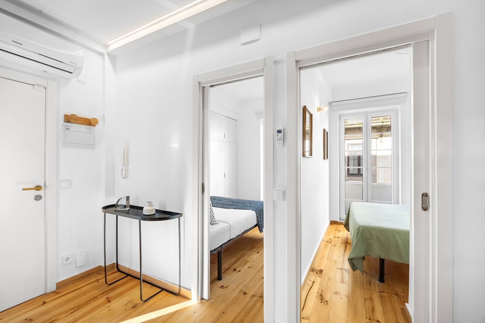 A hotel room with a wooden floor, a bed with a blue and white patterned bedspread, a small table with a black top, a white wall-mounted phone, and a white door with a keyhole.