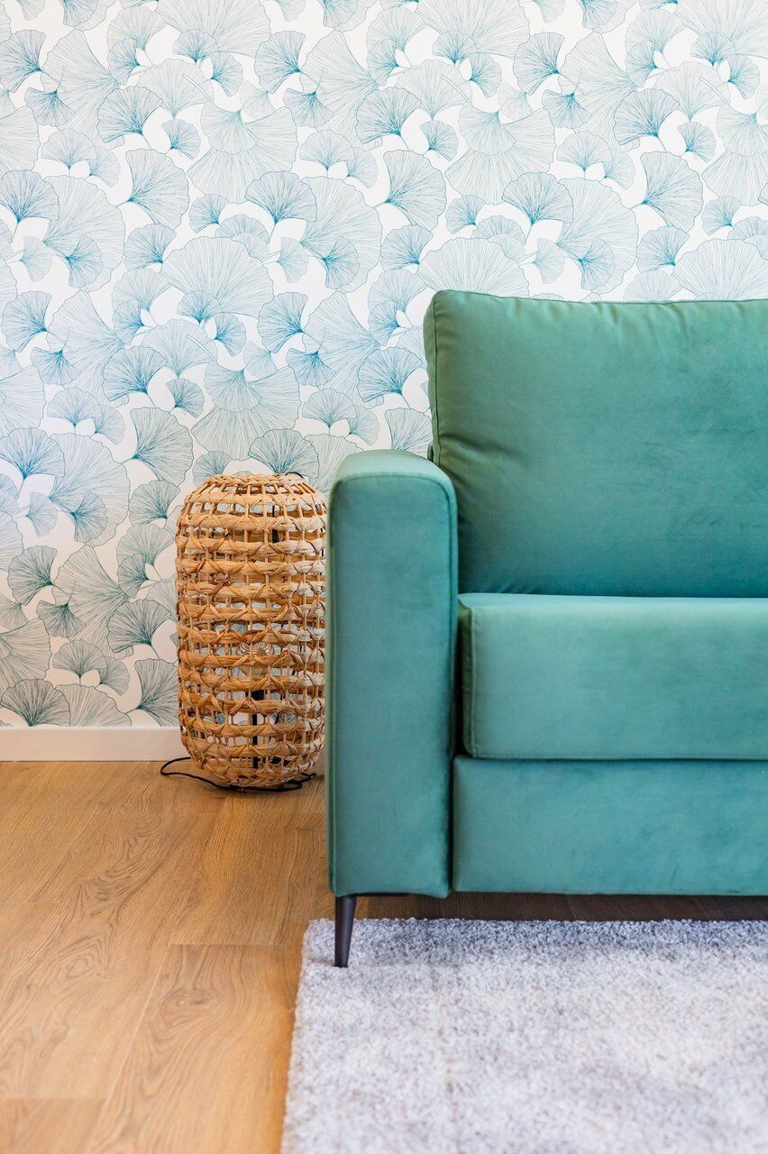 Un canapé de couleur turquoise, un tabouret en osier et un papier peint à motifs dans une pièce dotée d’un plancher en bois et d’un tapis blanc.