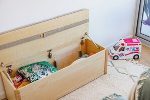 Un cofre de madera con tapa abatible, que contiene un cojín de estampado verde y blanco, un recipiente de plástico transparente y un coche de juguete de ambulancia.