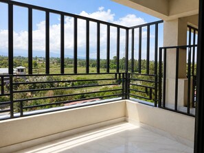 A balcony with a metal railing, a view of a green landscape, and a building in the distance.