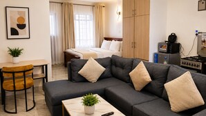 A modern hotel room with a sofa, a small table, a bed, a desk, and a kitchenette.