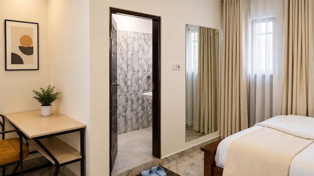 A hotel room with a bed, a desk, a chair, a potted plant, and a bathroom visible through an open door.