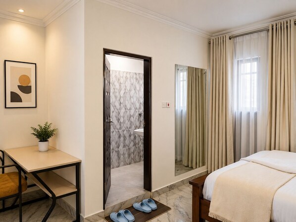 Habitación de hotel con cama, escritorio, silla, planta en maceta y baño visible a través de una puerta abierta.