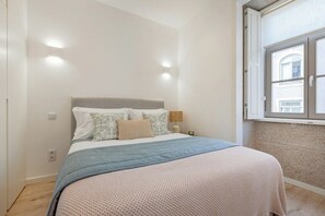 Ein Schlafzimmer mit einem Bett, Kissen, einem Nachttisch, einer Lampe und einem Fenster.