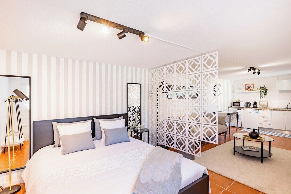 A modern hotel room with a bed, bedside table, lamp, and a view of the kitchen area.