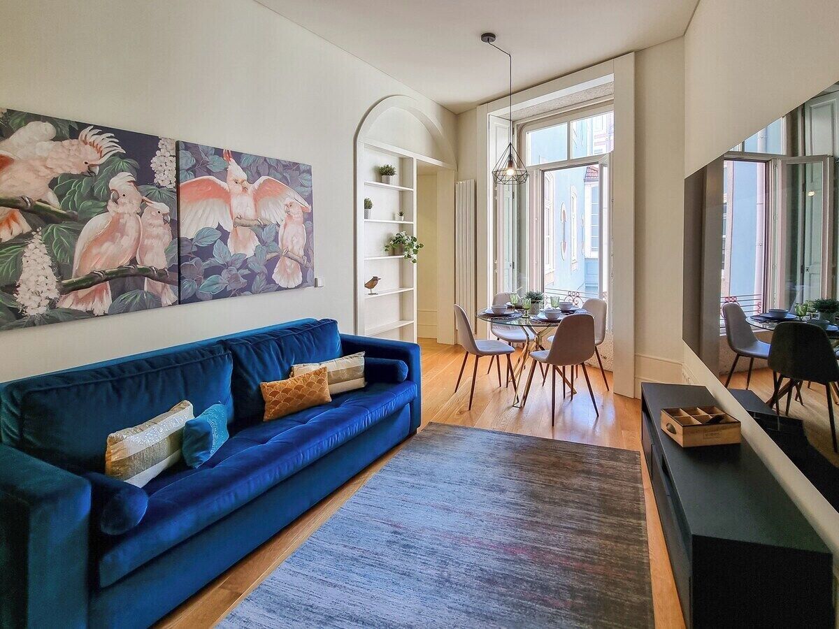 A living room with a blue sofa, a dining area with chairs, a bookshelf, and a painting of birds.
