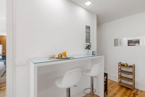 Eine moderne Küche mit weißen Wänden, einer Glas Arbeitsplatte, zwei weißen Barhocker und einem Holzregal mit Früchten.