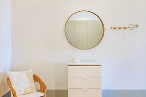 A room with a wicker chair, a white dresser, a round mirror, and a wall-mounted light fixture. - Boavista Light Studio (Porto)