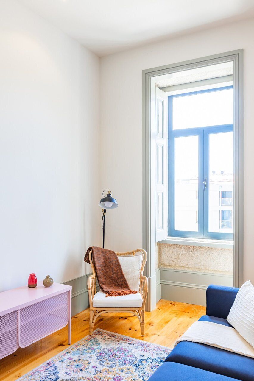 A room with a blue sofa, a wicker chair with a blanket, a pink console table, a floor lamp, and a large window with a view.