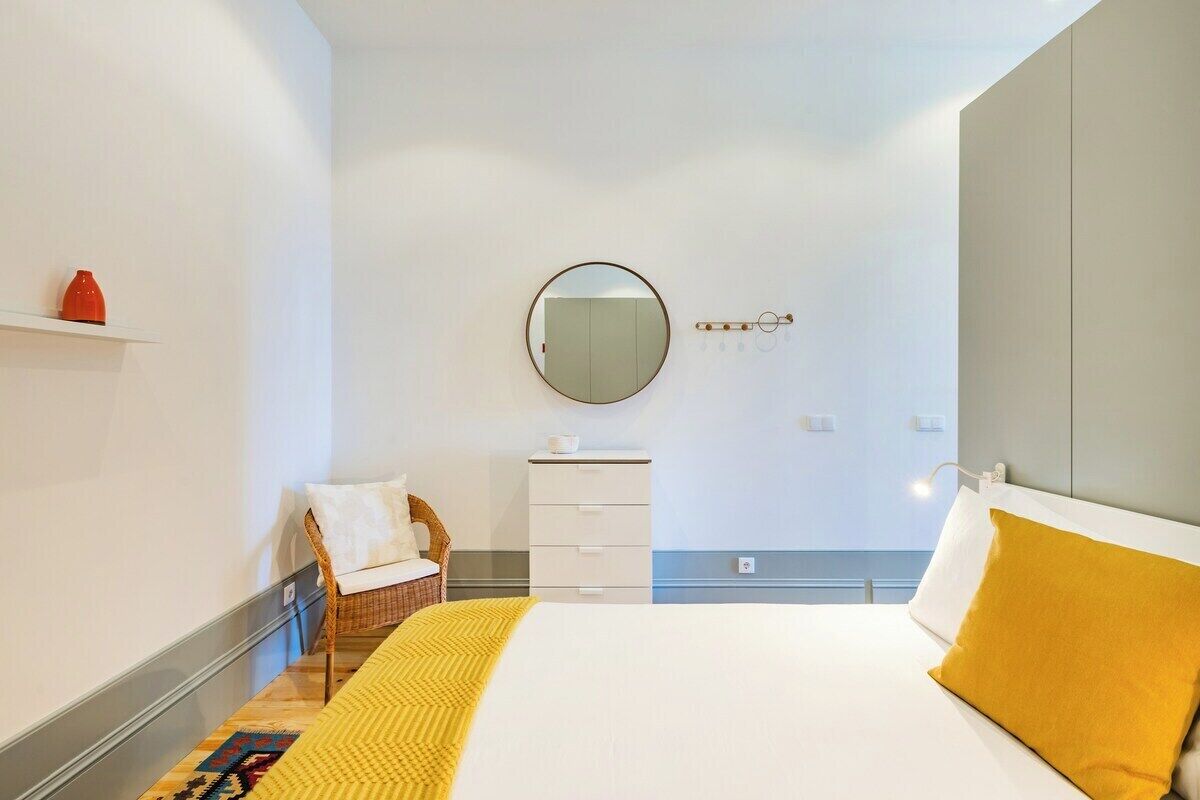 Ett hotellrum med en säng, en rund spegel, en liten vit byrå, en flätad stol och en hylla med en röd vas.