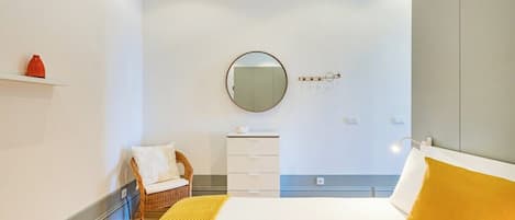 A hotel room with a bed, a round mirror, a small white dresser, a wicker chair, and a shelf with a red vase.
