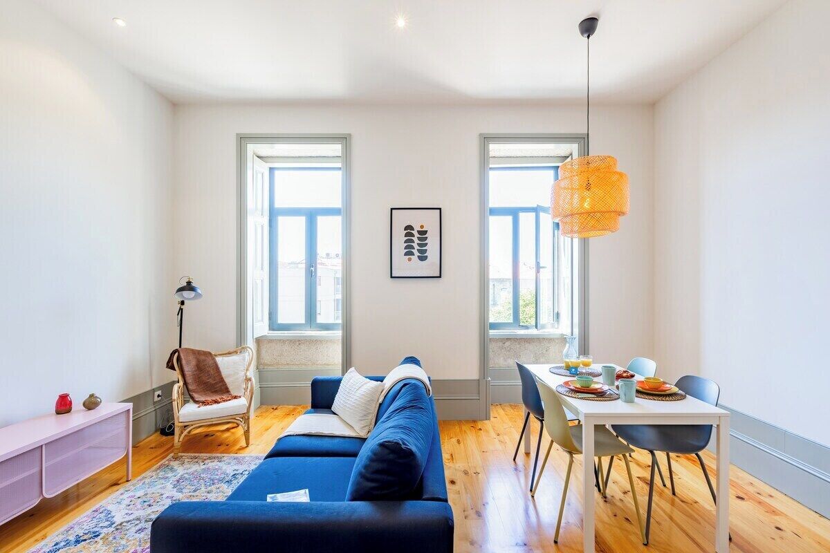 A modern living room with a blue sofa, a dining table with chairs, and two windows.