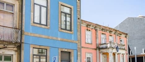 Una calle con coches aparcados, un edificio azul con una puerta y un edificio rosa con columnas.