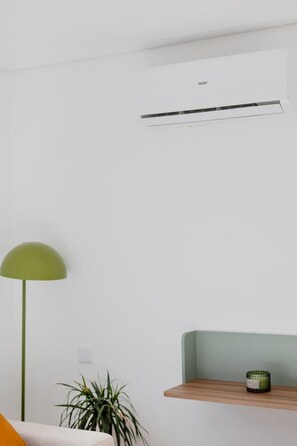 Ein Zimmer mit weißer Wand, einer grünen Stehlampe, einem Topfpflanze, einem hölzernen Regal mit einem grünen Behälter und einem weißen Klimagerät mit dem Aufdruck 'Haier'.