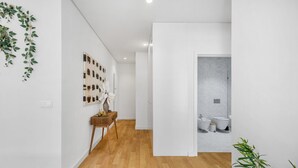 A hallway with a wooden floor, a white wall with a plant, a console table with decorative items, and a bathroom with a marble wall and toilet. - CharmINN Paraíso | Porto | AC (Porto)