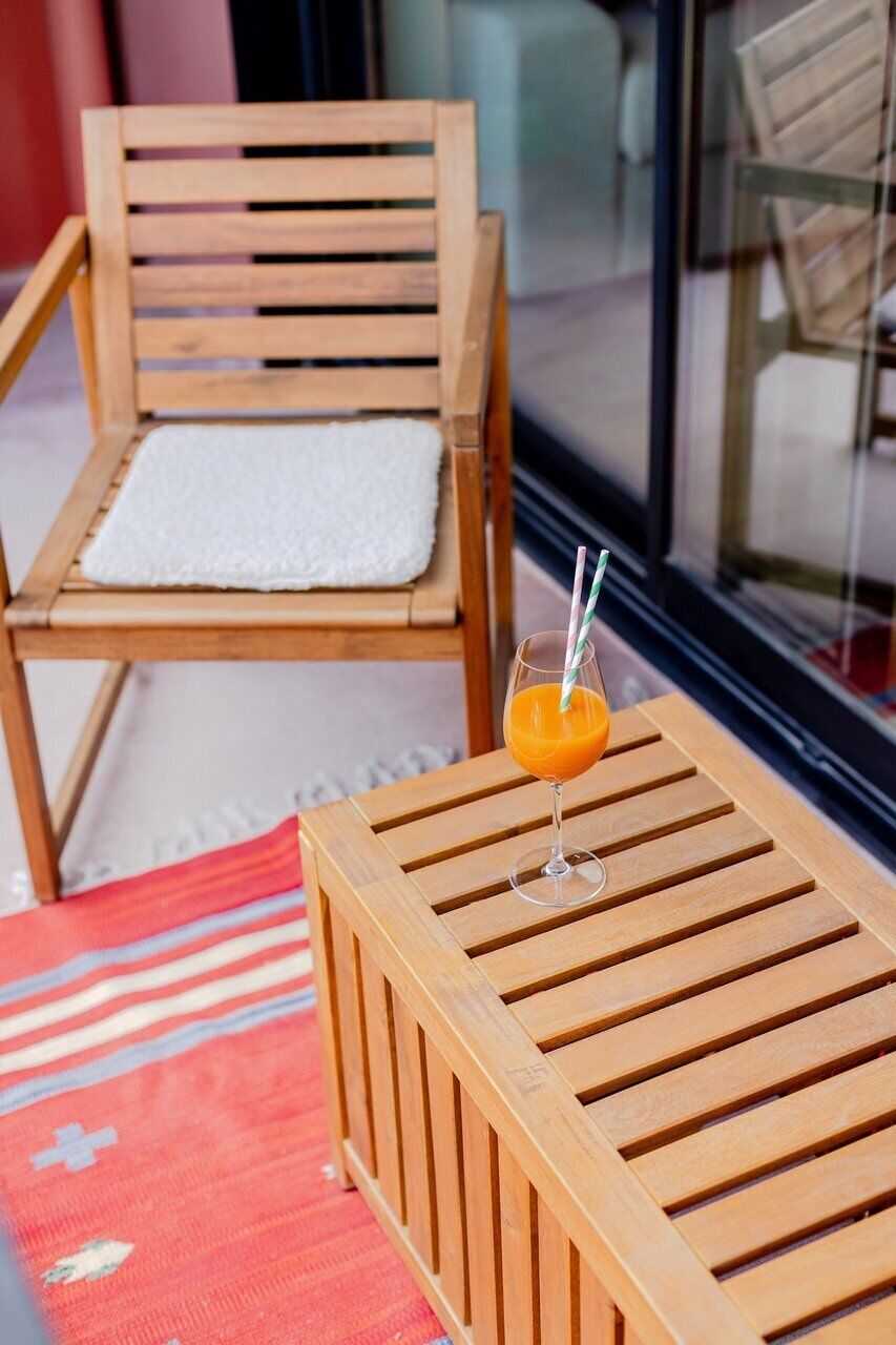 Ein Holzsessel mit weißem Kissen, ein Holztisch mit einem Glas Orangensaft und ein roter Teppich mit Muster.
