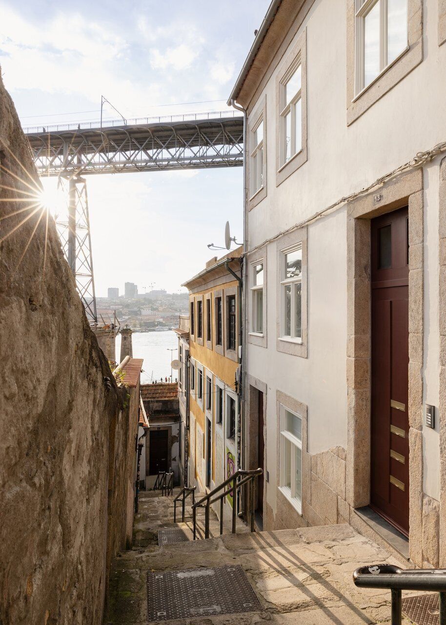 A narrow street with a bridge in the background, stone steps leading to a building with a red door, and a view of the water.