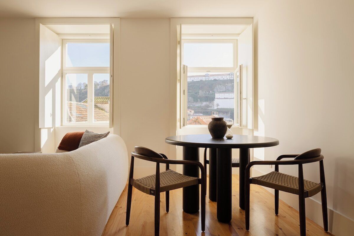 A room with a bed, a round table with chairs, and two windows with a view of a cityscape.