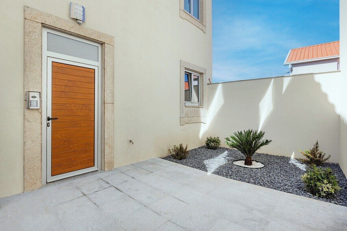 Un edificio moderno con una puerta de madera, una ventana pequeña y un área ajardinada con plantas y grava.