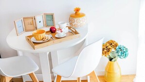 Uma mesa branca com um tabuleiro, quadros enquadrados, um vaso amarelo com flores e cadeiras brancas.