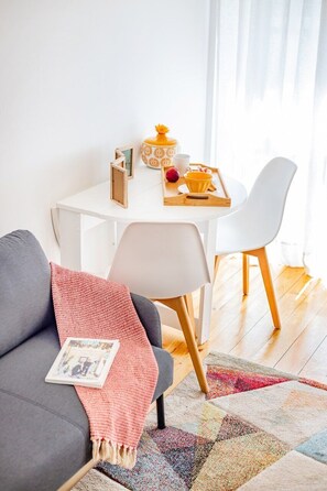 Ein gemütliches Wohnzimmer mit einem grauen Sofa, einem weißen Tisch mit einer Speiseplatte, einem gelben Dekoartikel und einem bunten Teppich.