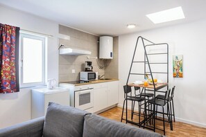 Eine kompakte Küche mit Mikrowelle, Wasserkocher und Kaffeemaschine, ein kleiner Essbereich mit Tisch und Stühlen, eine Couch und ein Fenster mit Vorhängen.