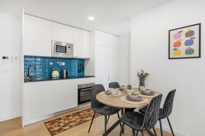Une cuisine compacte avec des meubles blancs, un pare-feu bleu et un four à micro-ondes. On y trouve un coin repas avec une table ronde et des chaises, ainsi qu’une œuvre d’art encadrée accrochée au mur.