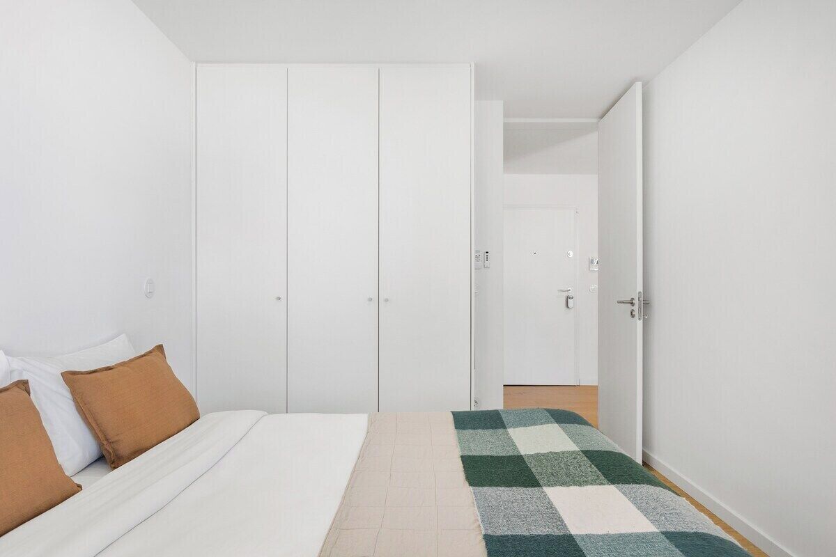 Une chambre à coucher avec un lit, deux oreillers marron, une couverture à carreaux, une armoire et une porte donnant sur une autre pièce.